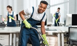 cheerful african american cleaner vacuuming floor while looking at camera